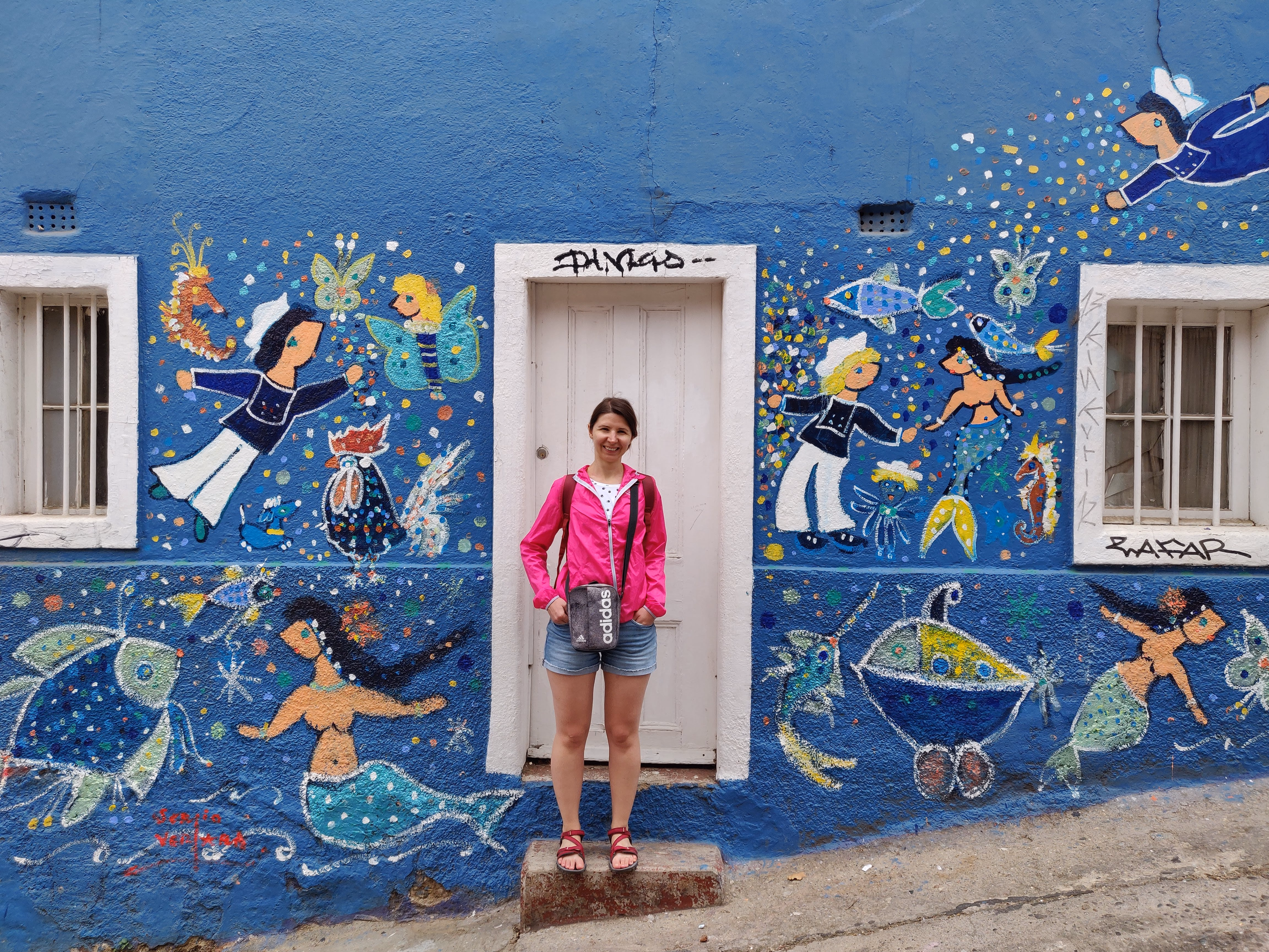 A cobalt-blue wall covered in nautical folk art — flying sailors, mermaids, seahorses and fish painted around a white door on a steep lane.