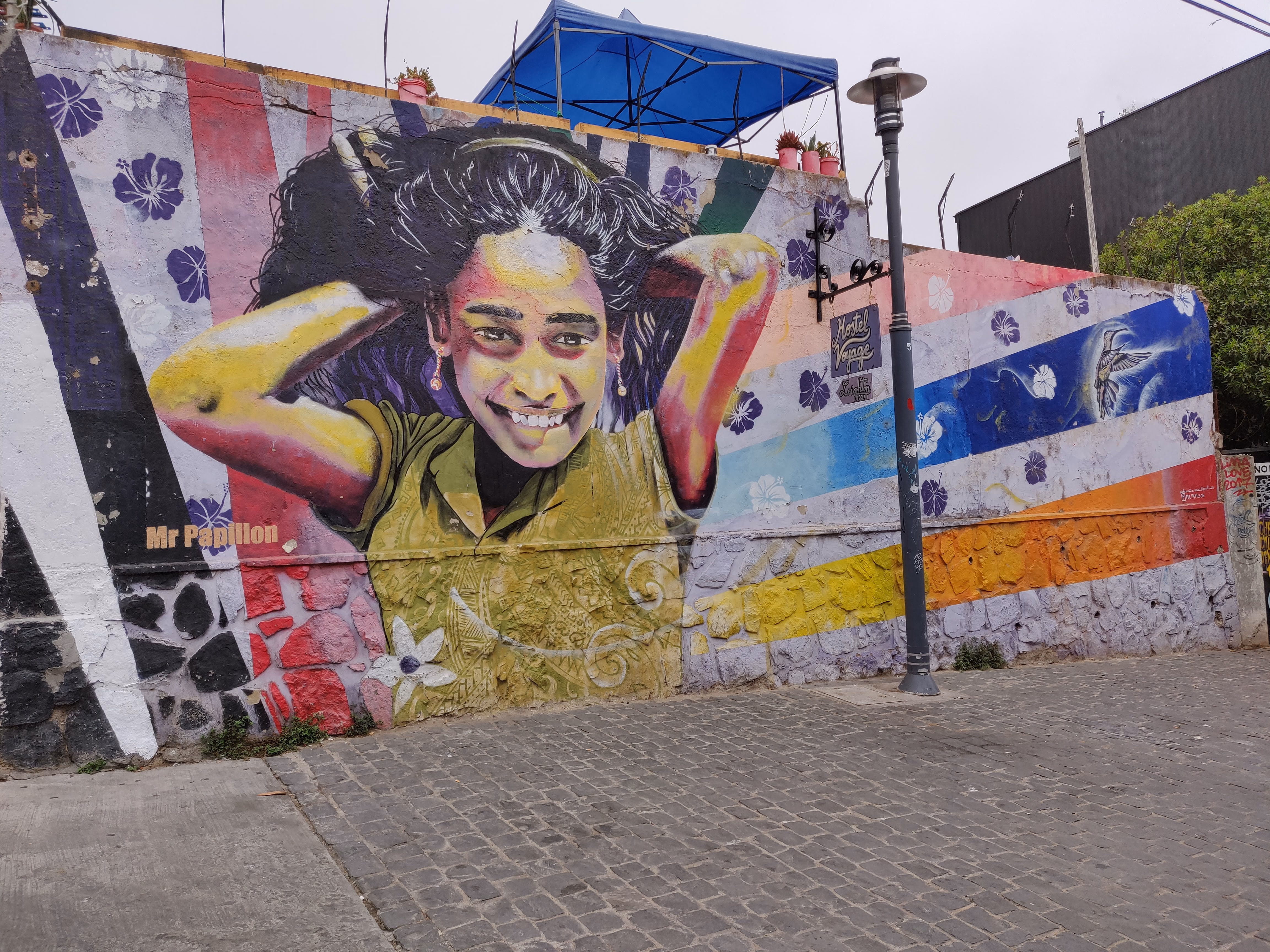 A smiling girl with hands in her hair dominates a long wall on cobblestones — purple hibiscus, striped panels and a hummingbird beside a Hostel Voyage sign.