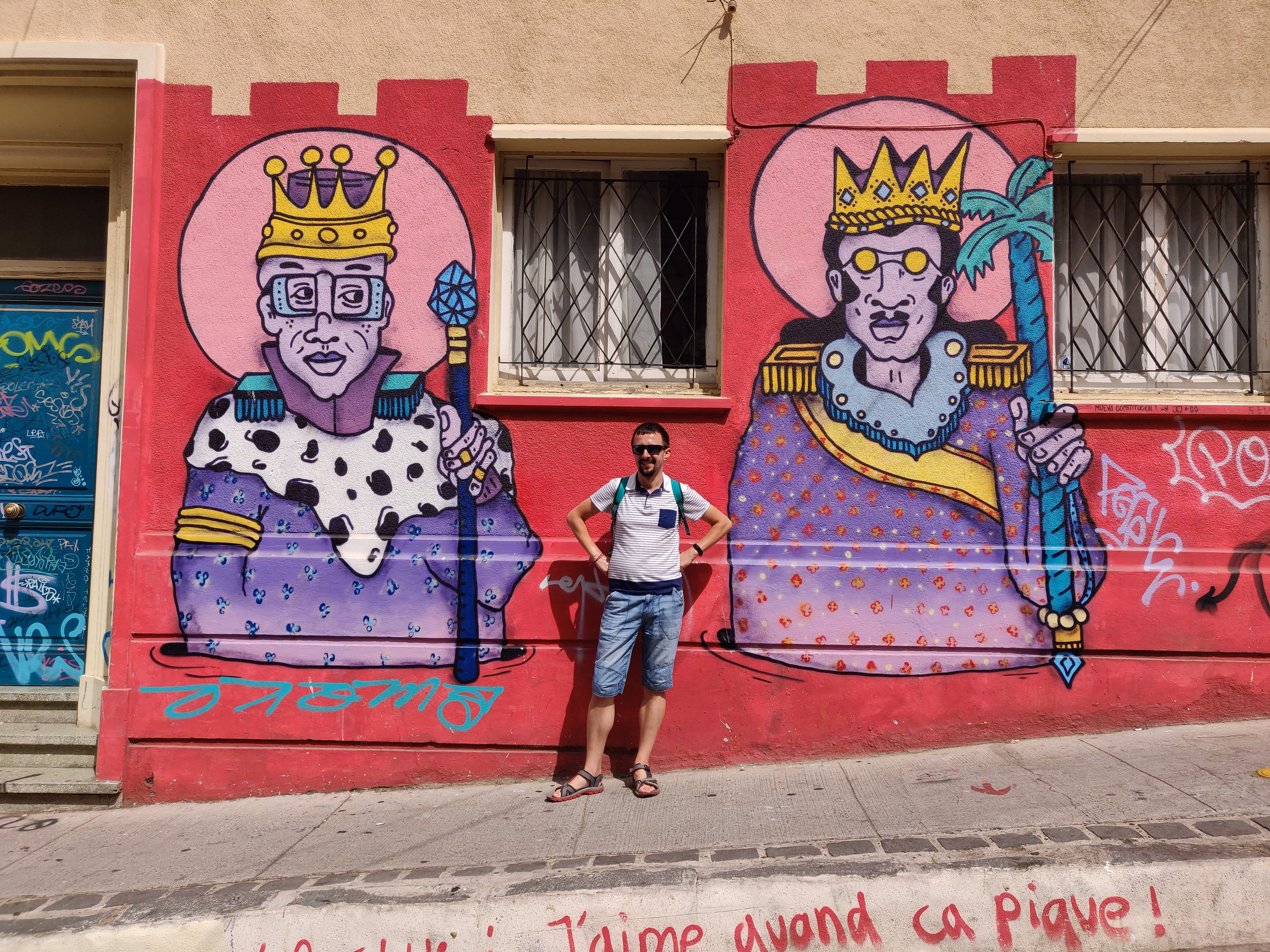 Two crowned figures in purple robes and pink halos flank a doorway on a fuchsia wall — crenellated battlements painted above barred windows.