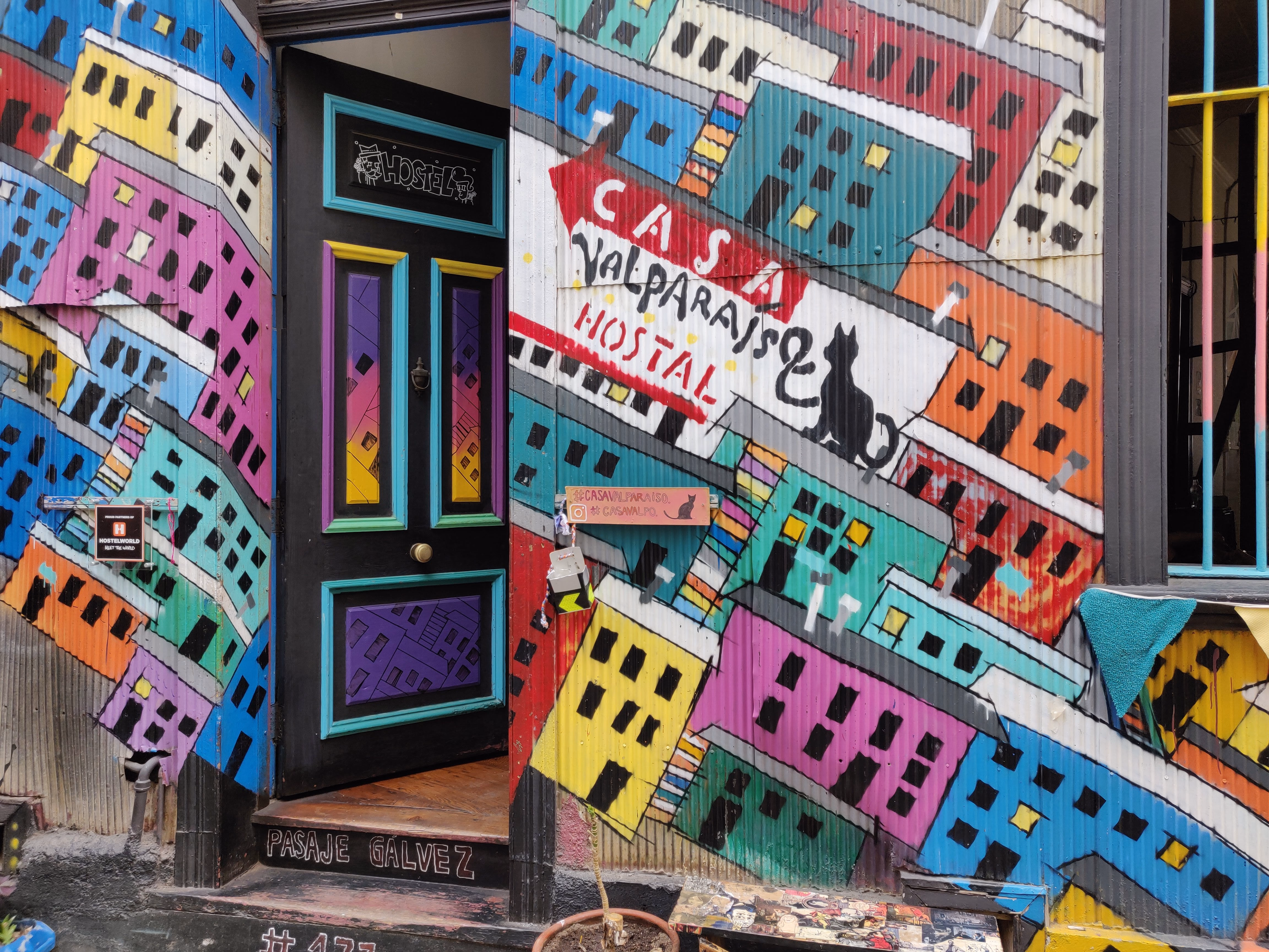 A hostel entrance painted as a hillside of stacked yellow, blue and pink houses — "Casa Valparaíso Hostal" in red lettering beside a black cat silhouette.