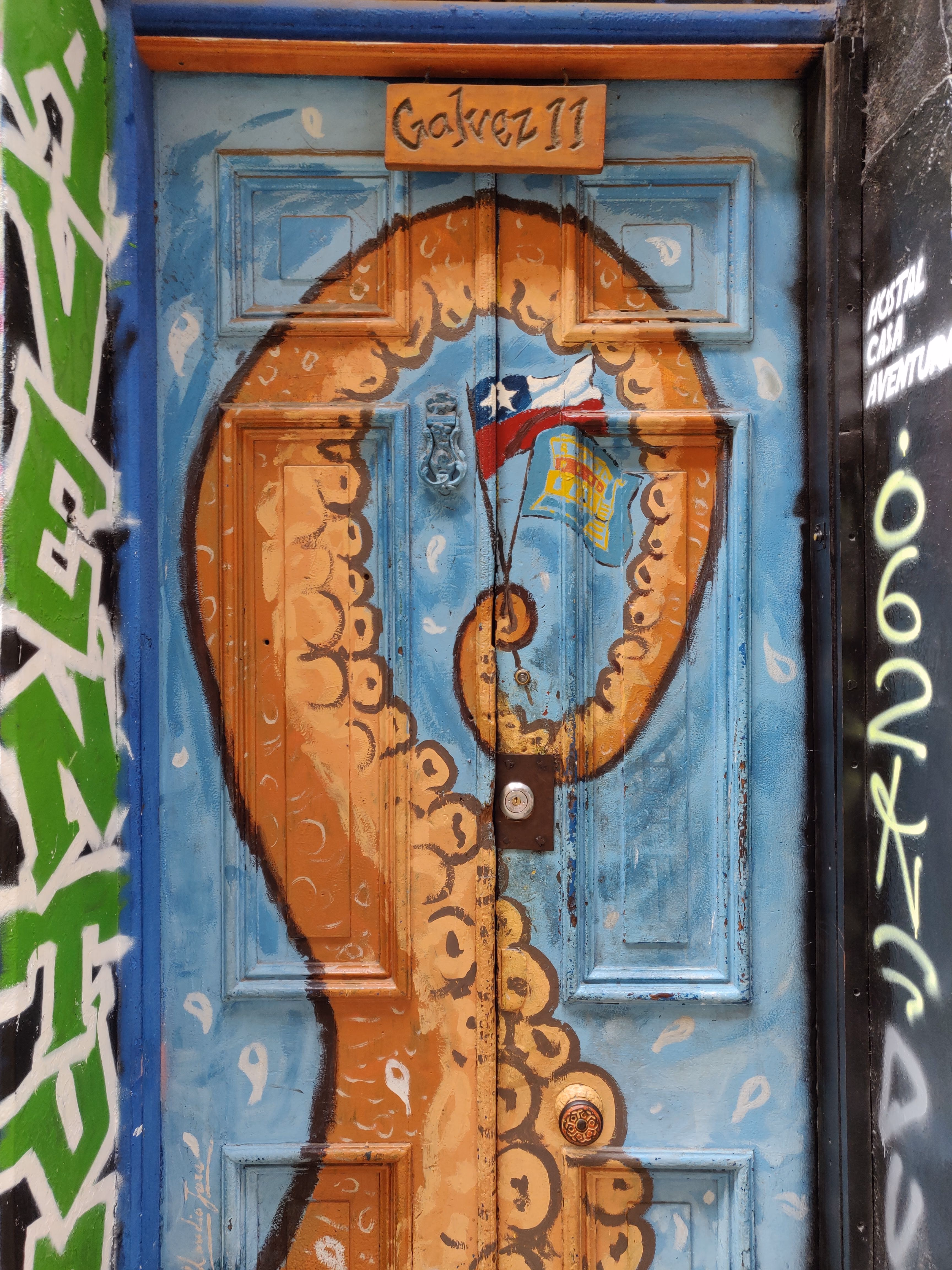 An orange octopus tentacle curls across a sky-blue door scattered with white teardrops — a Chilean flag painted at the centre, "Gálvez 11" on a wooden sign above.