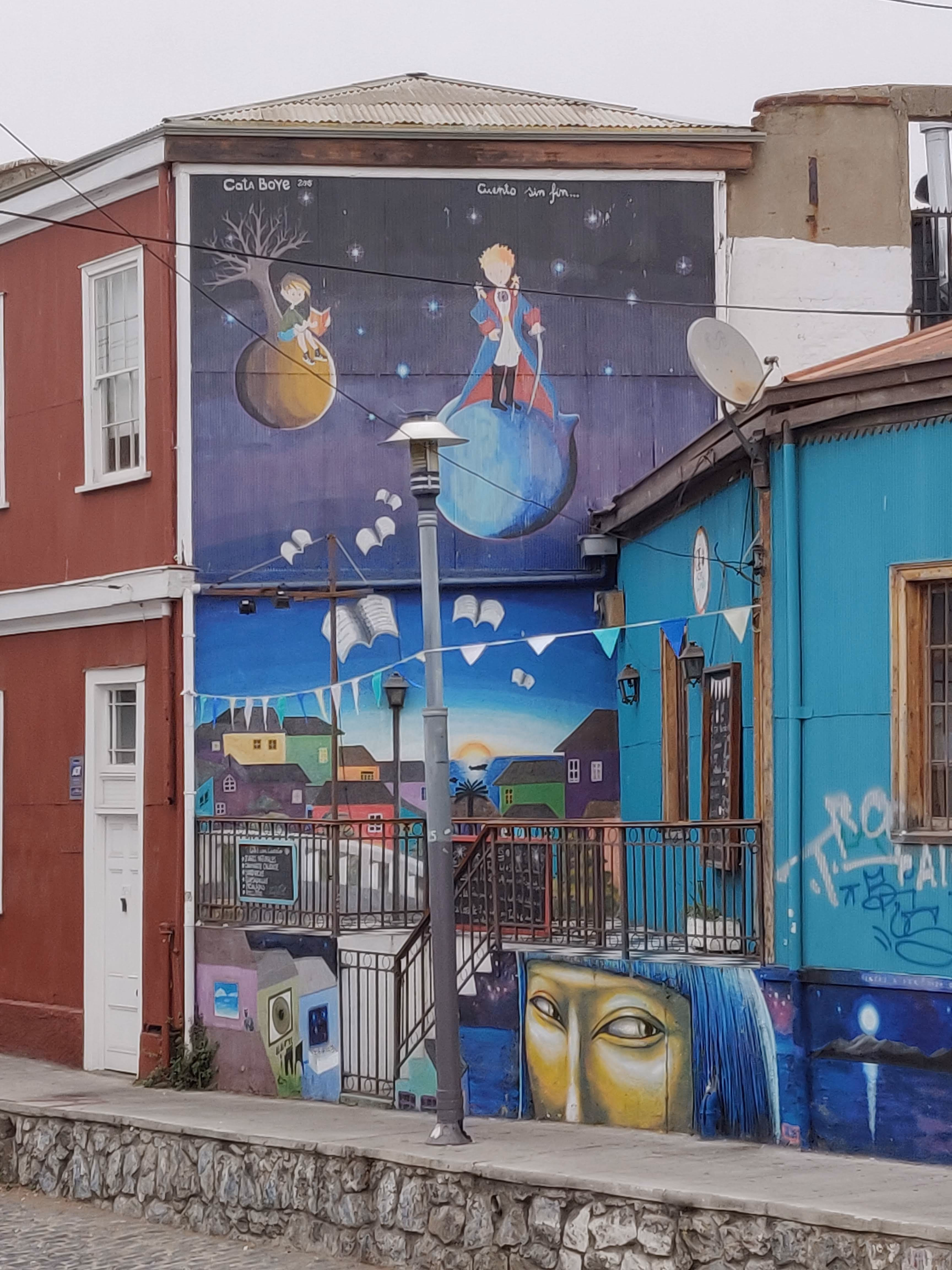 A three-storey mural — the Little Prince on his planet above colourful cerro houses, an open book floating in the sky, a golden face with blue hair below a terrace railing.