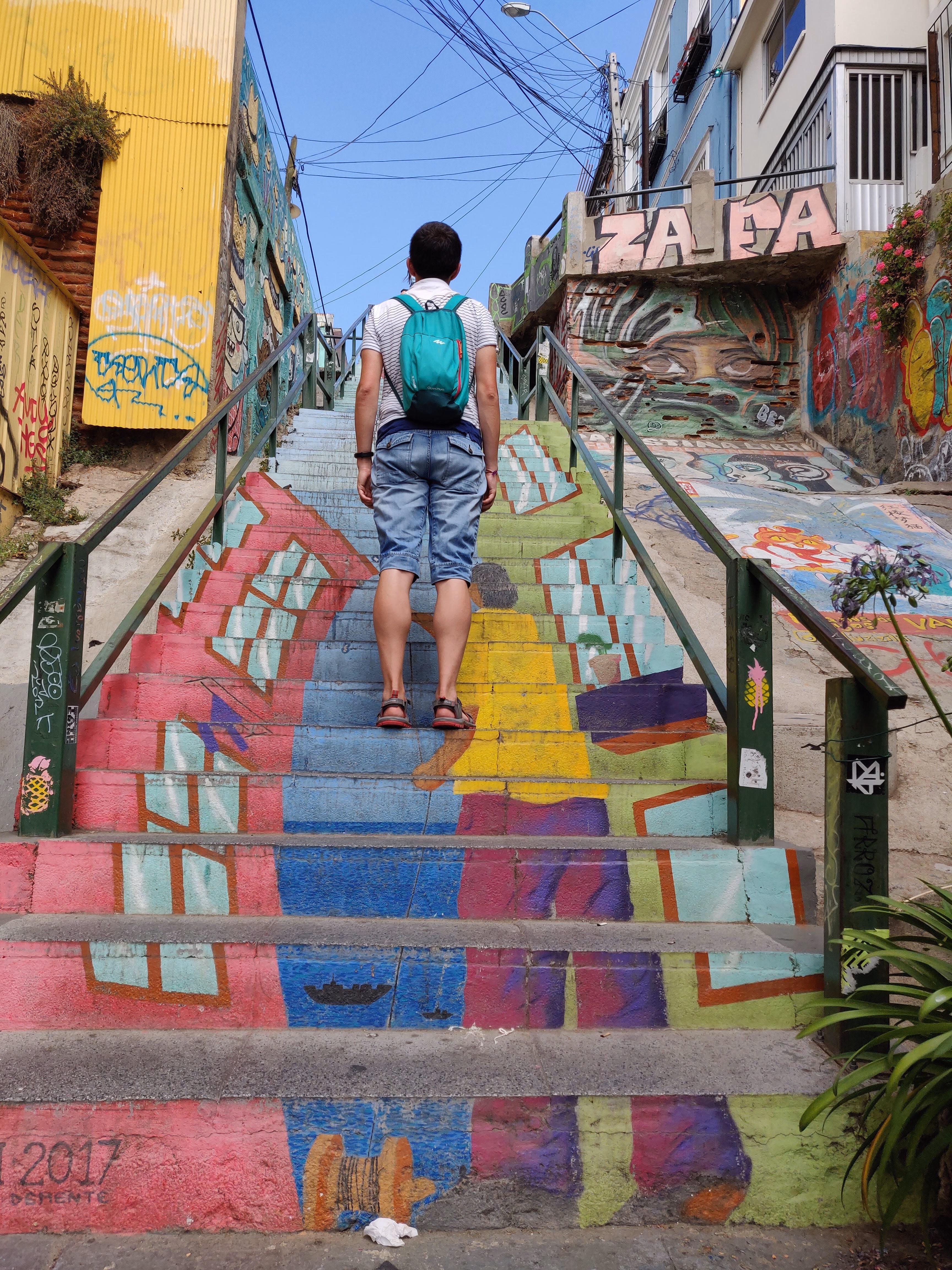 A figure in a yellow shirt climbs a wide staircase painted as a hillside of pink, blue and yellow houses — green handrails tagged on both sides.
